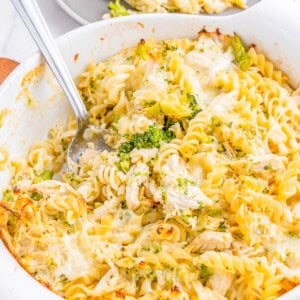 A white casserole dish filled with baked pasta, broccoli, and chicken, served with a spoon. Another portion is plated in the background on a gray plate.
