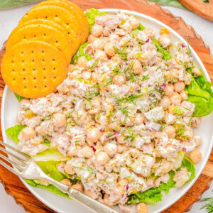 A plate of chickpea salad with visible ingredients including chickpeas, herbs, and dressing, garnished with fresh dill. The plate is accompanied by three round crackers and a fork, placed on a wooden board.