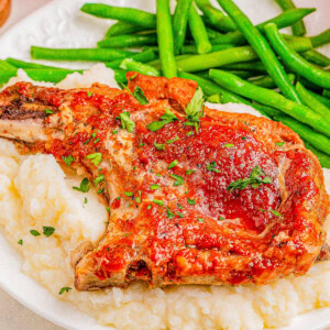A plate with a pork chop garnished with herbs on a bed of mashed potatoes, accompanied by green beans.