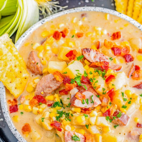 A bowl of creamy corn chowder with sausage pieces and diced potatoes, garnished with chopped parsley and crumbled bacon. Corn on the cob and saltine crackers are placed beside the bowl.