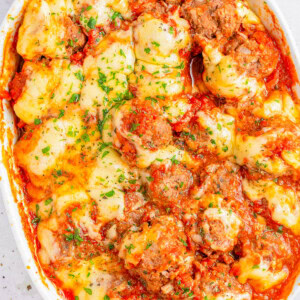 A baked dish in a white casserole pan featuring meatballs, melted cheese, and tomato sauce garnished with chopped parsley. A serving spoon is partially visible on the right side of the dish.