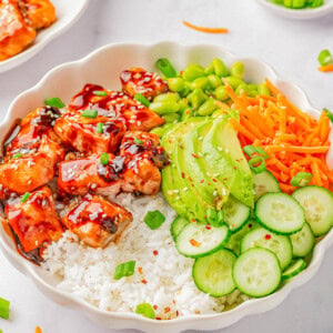 A bowl of rice topped with glazed salmon, sliced avocado, shredded carrots, sliced cucumber, edamame, and garnished with chopped green onions and sesame seeds.
