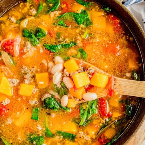 A pot of soup containing chunks of squash, white beans, spinach, tomatoes, and ground meat, with a wooden spoon scooping some out.
