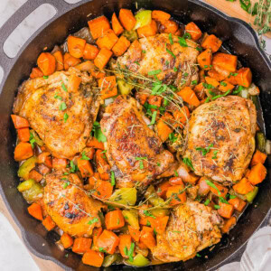 A cast-iron skillet filled with cooked chicken thighs, diced sweet potatoes, and green bell peppers, garnished with chopped parsley. A cutting board with chopped parsley and a bowl of salt are beside the skillet.