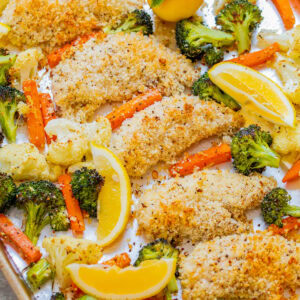 Sheet pan meal of lemon-baked chicken with breaded coating, surrounded by roasted vegetables including broccoli, carrots, and cauliflower, garnished with lemon wedges.