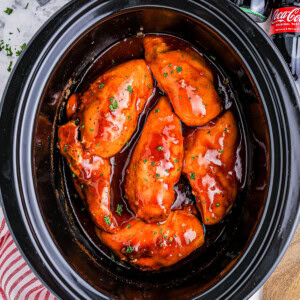 Slow cooker with five pieces of chicken breast coated in a red sauce, on a countertop with a striped towel, wooden spoon, salt and pepper shakers, and two cola bottles in the background.