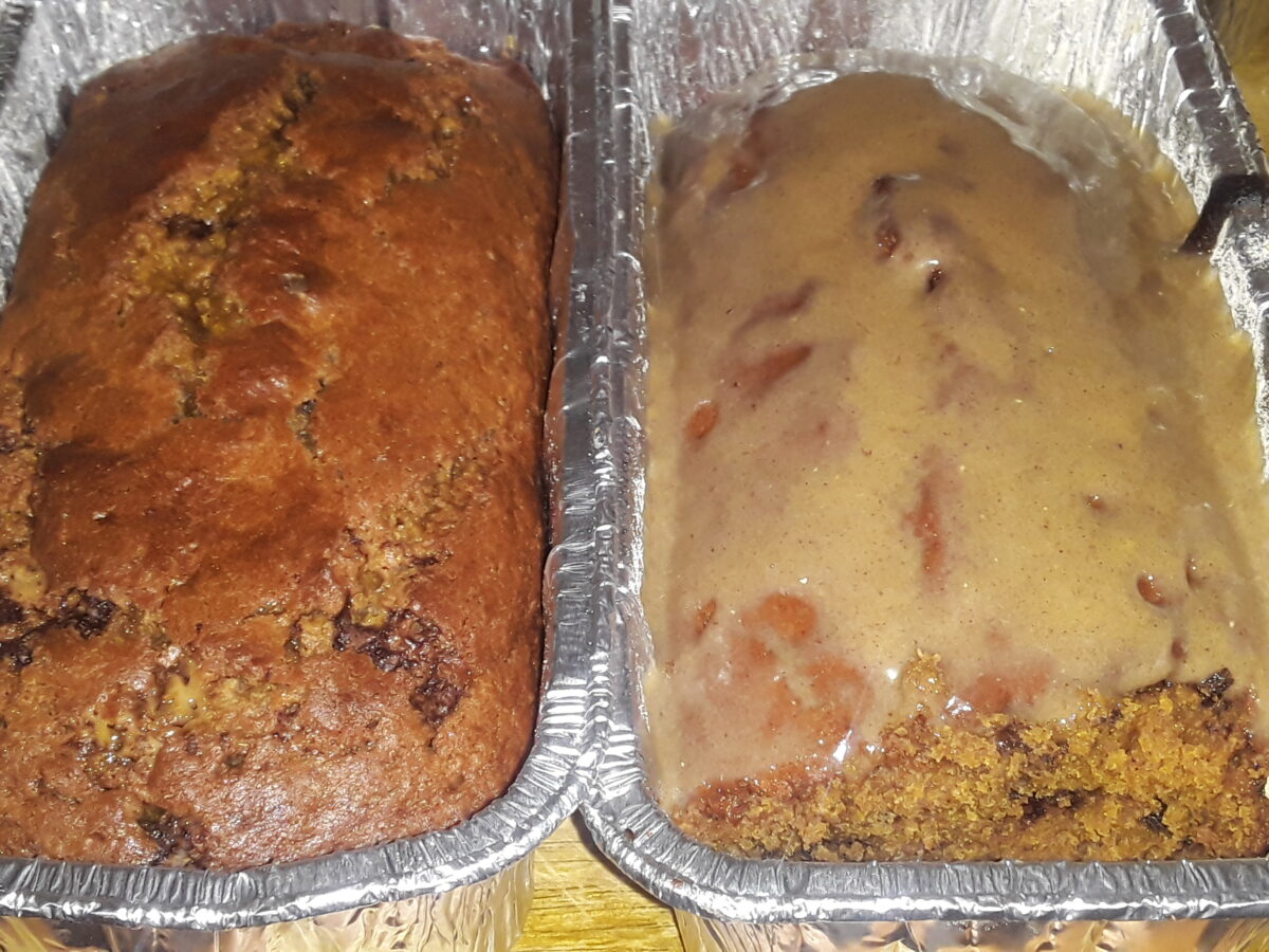 Two loaves of bread in foil trays on a wooden surface; the left loaf is darker and crisper, while the right has a glossy brown glaze.