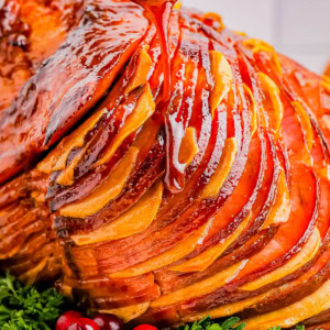 A glazed ham with sliced sections is displayed on a white plate, garnished with fresh cranberries and herbs, as honey is drizzled over the top.