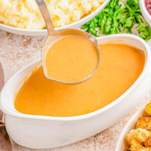 A ladle dips into a gravy boat filled with thick, brown gravy. Surrounding it are bowls of mashed potatoes and stuffing, garnished with parsley.