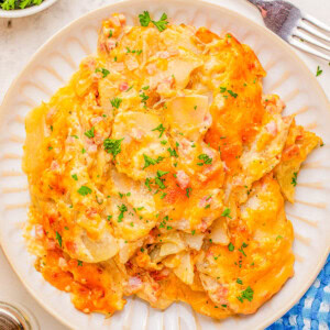 A plate of cheesy scalloped potatoes garnished with parsley, next to a fork, a blue cloth, and a small dish of fresh herbs.