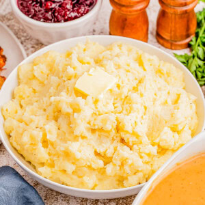 A bowl of mashed potatoes with butter on top, surrounded by bowls of cranberry sauce and gravy, with salt and pepper shakers in the background.