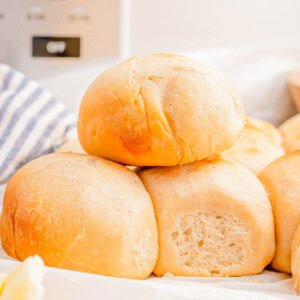 A stack of freshly baked dinner rolls on a table with a knife and butter in the foreground.