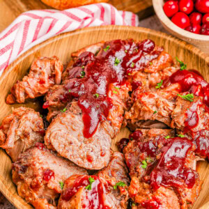 Sliced pork tenderloin topped with cranberry sauce in a wooden bowl, accompanied by a bowl of cranberries and a loaf of bread.