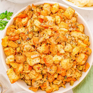 A bowl of stuffing made with croutons, herbs, and vegetables is placed on a table. Other dishes, including gravy and mashed potatoes, are partially visible nearby.