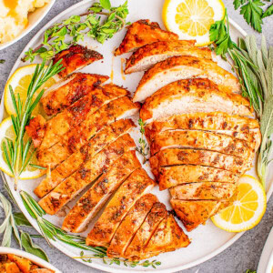 Sliced roasted turkey breast on a plate with lemon slices and herbs, surrounded by dishes of mashed potatoes and stuffing.