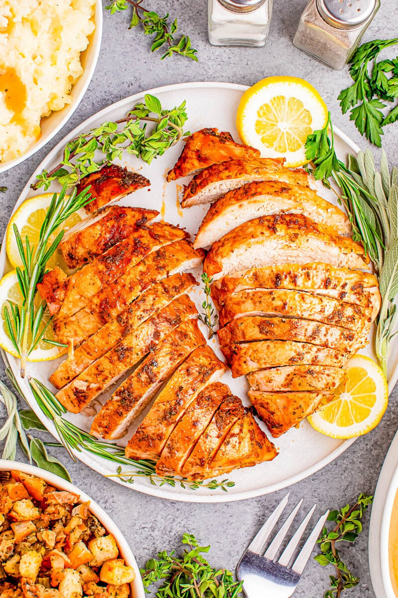 Sliced roasted turkey breast on a plate with lemon slices and herbs, surrounded by dishes of mashed potatoes and stuffing.