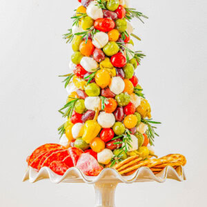 A tree-shaped appetizer display featuring grape tomatoes, mozzarella balls, olives, and peppers on skewers, garnished with rosemary, with salami and crackers at the base on a pedestal stand.