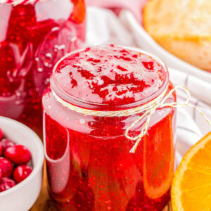Jar of red jam next to fresh cranberries and an orange slice on a wooden surface, with a larger jar of jam and pastries in the background.
