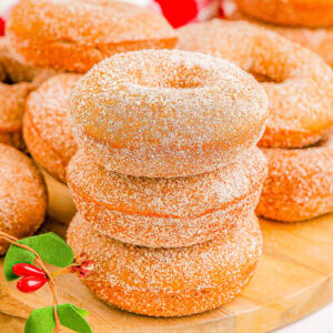 A stack of three sugar-coated donuts on a wooden board, surrounded by more donuts and sprigs with red berries.