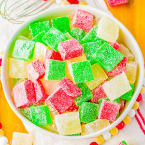 A bowl of colorful, sugar-coated gumdrops in red, green, and yellow on a wooden tray with a whisk nearby.