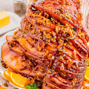 A spiral-cut ham glazed with honey and pecan topping, garnished with orange slices and parsley on a white plate. Butter and condiments are in the background.