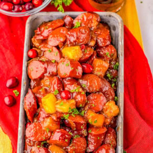 Rectangular dish with glazed sausage slices and pineapple chunks, garnished with parsley. A bowl of cranberries and a glass of iced drink are in the background, on a red cloth.