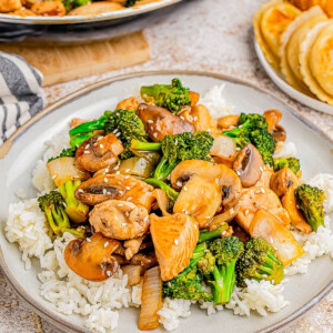 A plate of stir-fried chicken, mushrooms, and broccoli served over white rice. Nearby, there is a plate with dumplings and fried chicken.