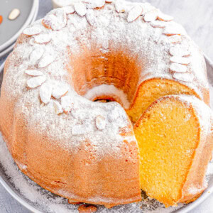 Bundt cake dusted with powdered sugar and topped with almond slices on a gray plate, featuring a single slice cut out; surrounded by scattered almonds and small dishes.