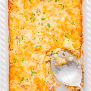 A baked casserole in a white dish, topped with melted cheese and garnished with herbs. A large serving spoon rests in an empty section.