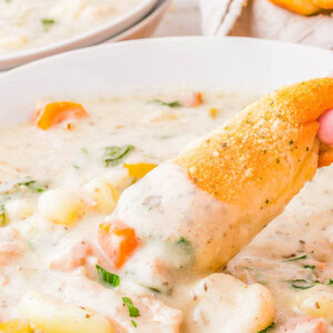 A bowl of creamy chicken gnocchi soup with visible chunks of chicken, carrots, and spinach. A hand dips a piece of bread into the soup. Another bowl and bread are blurred in the background.