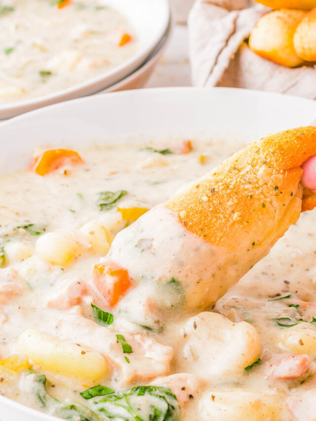 A bowl of creamy chicken gnocchi soup with visible chunks of chicken, carrots, and spinach. A hand dips a piece of bread into the soup. Another bowl and bread are blurred in the background.