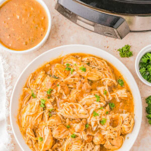 A bowl of shredded chicken in sauce, garnished with herbs, next to a slow cooker and a small bowl of extra sauce on a marble surface.
