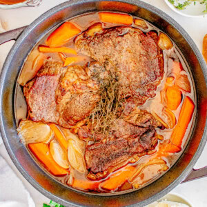 A stew pot with cooked beef, carrots, and onions, garnished with herbs, surrounded by ingredients like broth, herbs, and blue cheese on a white surface.