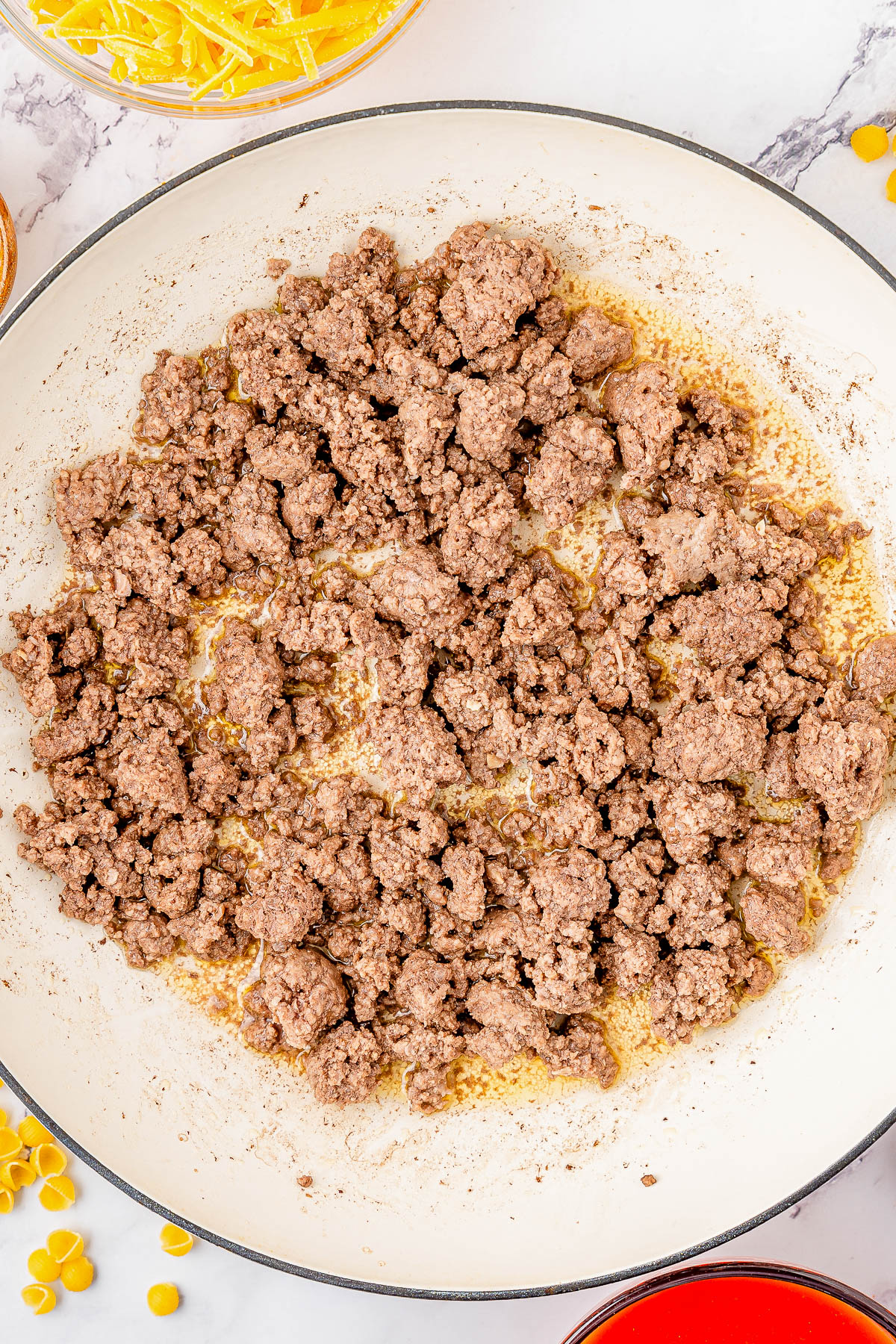 Ground beef cooking in a white skillet on a marble countertop, surrounded by bowls of grated cheese and yellow ingredients.
