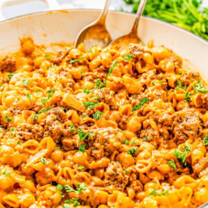 A white skillet filled with cheesy pasta, ground beef, and garnished with chopped parsley. Two serving spoons rest in the dish. Parsley is visible in the background.