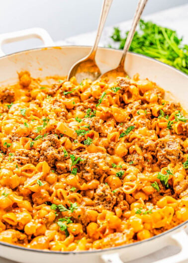 A white skillet filled with cheesy pasta, ground beef, and garnished with chopped parsley. Two serving spoons rest in the dish. Parsley is visible in the background.