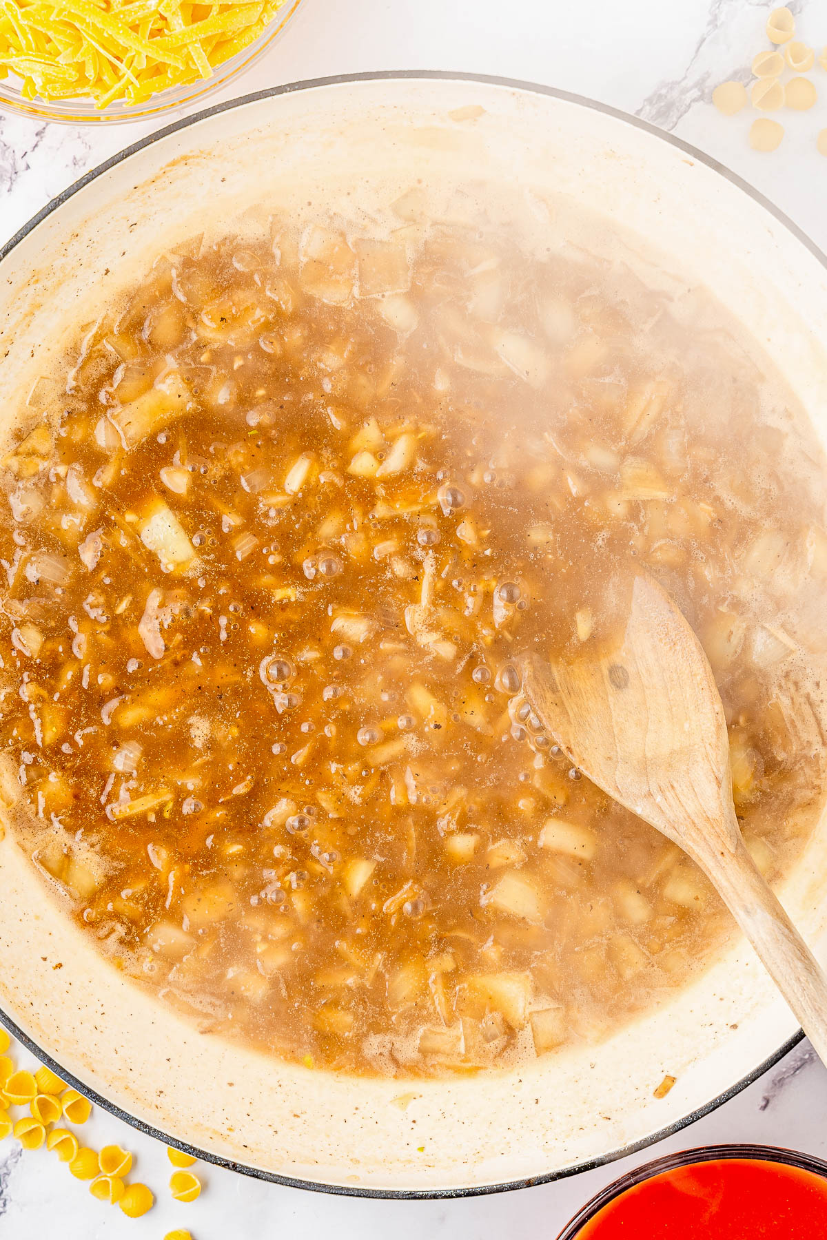 A pot of onion soup being simmered, with a wooden spoon stirring the mixture. Steam is rising from the surface, and uncooked pasta is visible in the corner.
