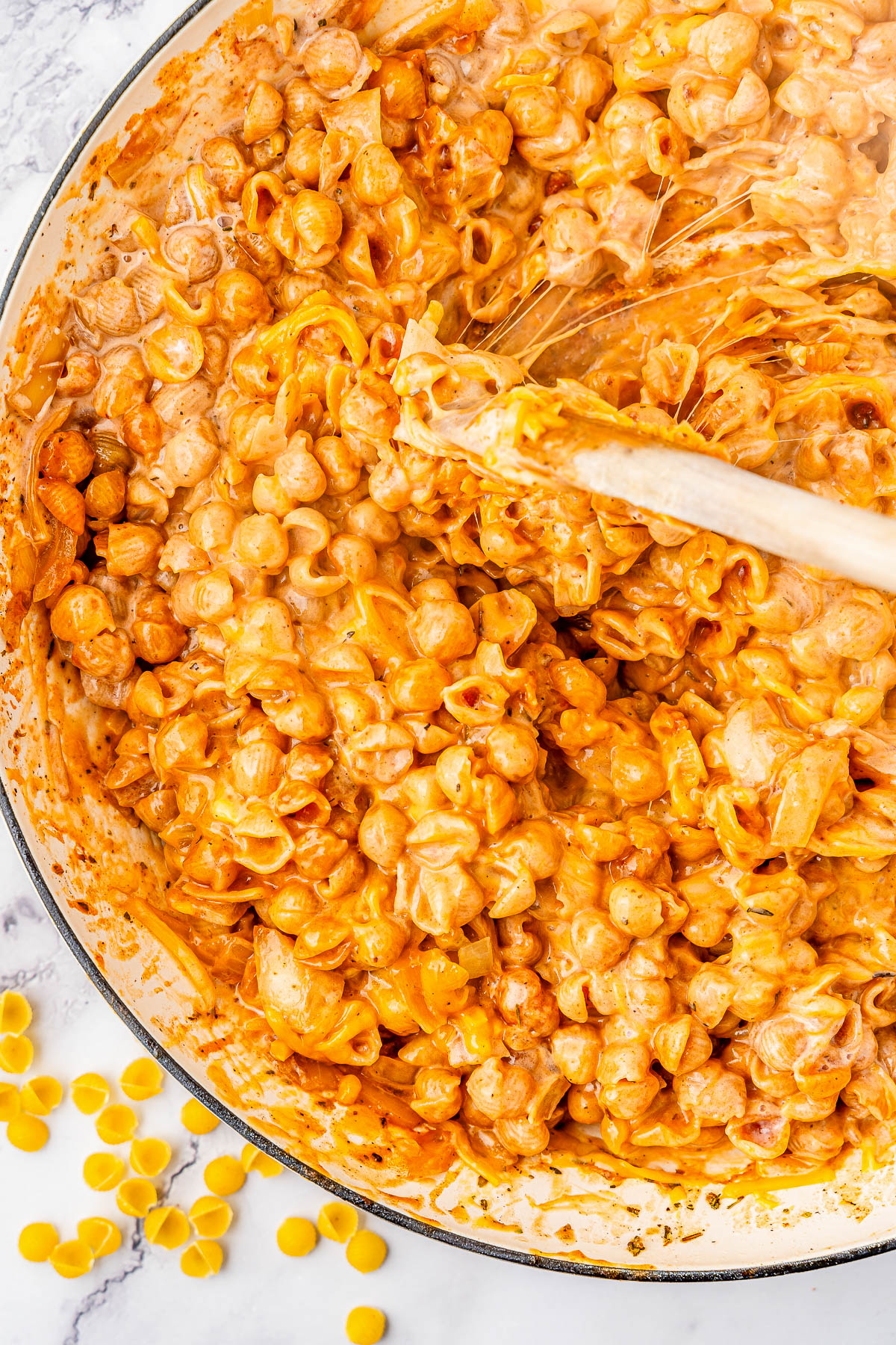 A pot filled with creamy pasta shells in a rich, orange-colored sauce, with a wooden spoon stirring the mixture. Some uncooked pasta shells are scattered nearby on a marble surface.