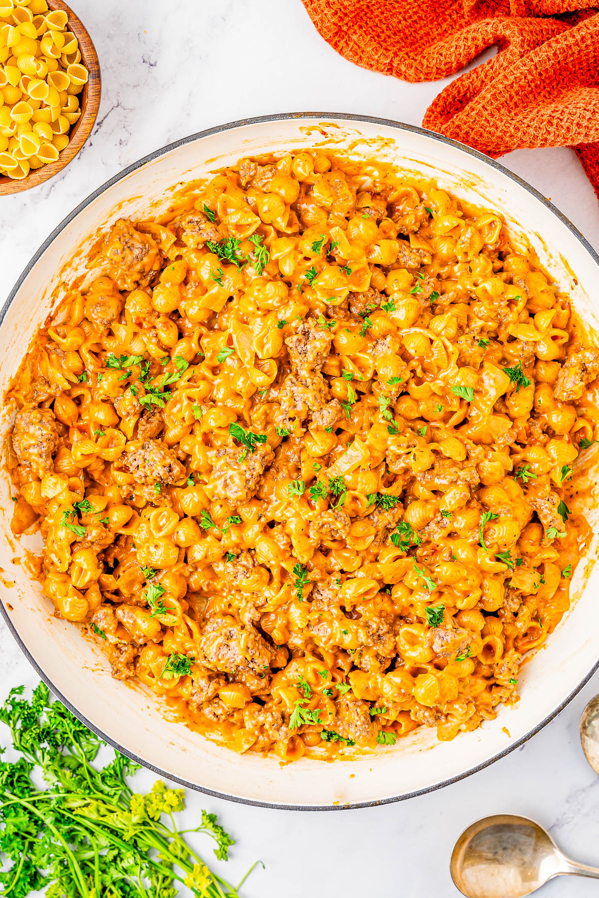 A large pan filled with creamy pasta mixed with ground meat and garnished with parsley. Spoons, uncooked pasta, and a red cloth are nearby.