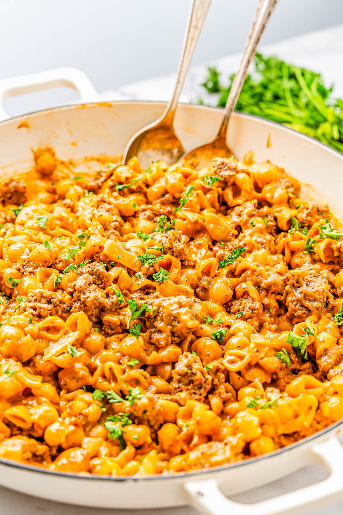 A skillet filled with creamy pasta and ground meat garnished with fresh parsley, with two serving spoons.