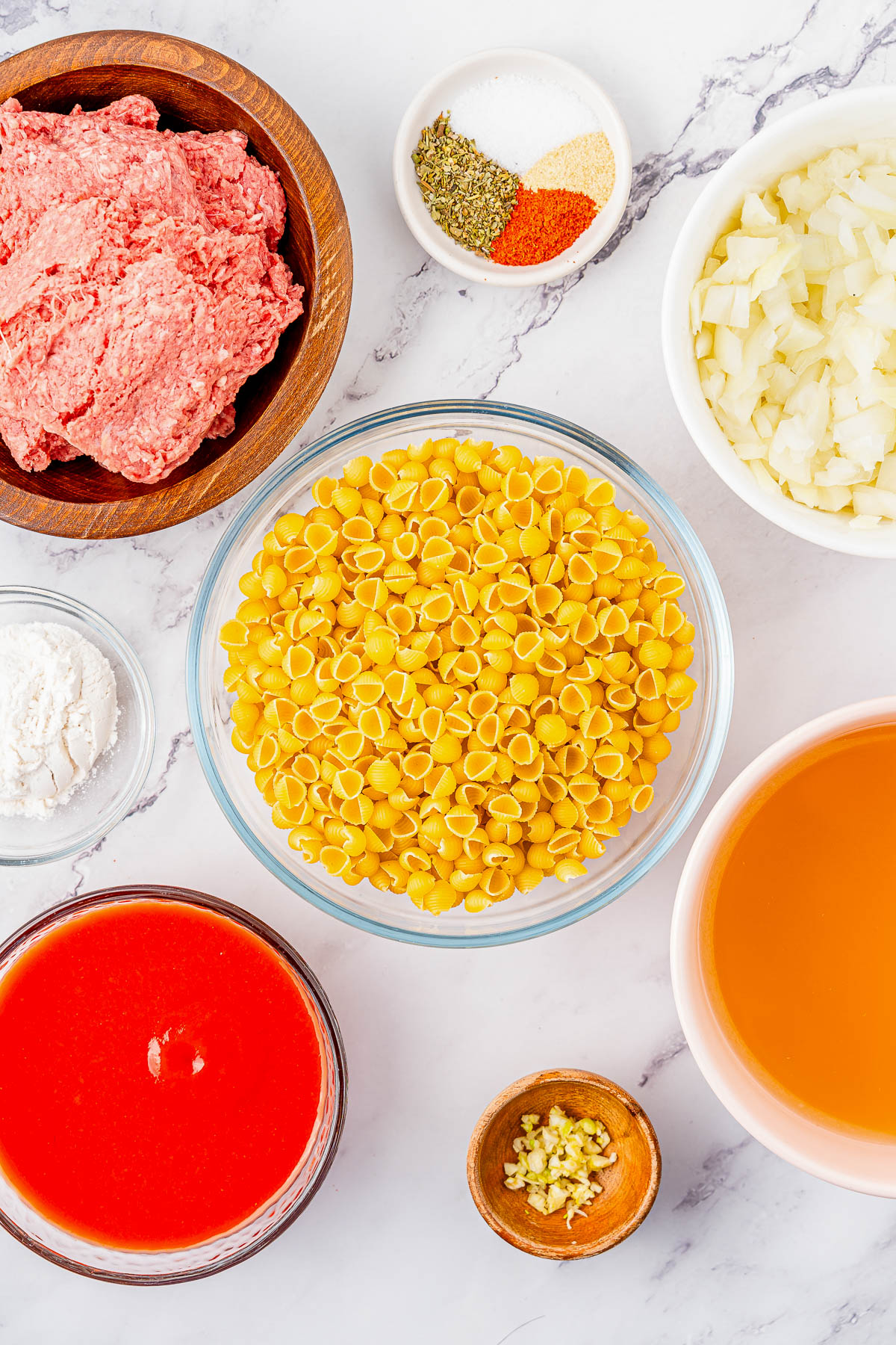 Ingredients on a marble counter: ground meat, pasta shells, chopped onions, garlic, tomato sauce, broth, spices, and flour.