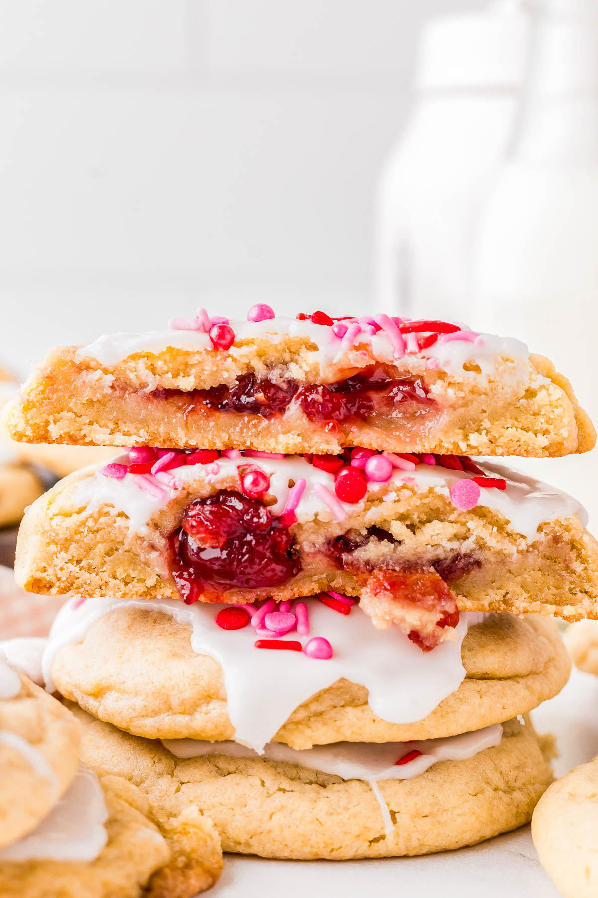 Stacked cookies with white icing and red sprinkles; a cookie is halved, revealing a red, jam-like filling. Blurred milk bottles in the background.