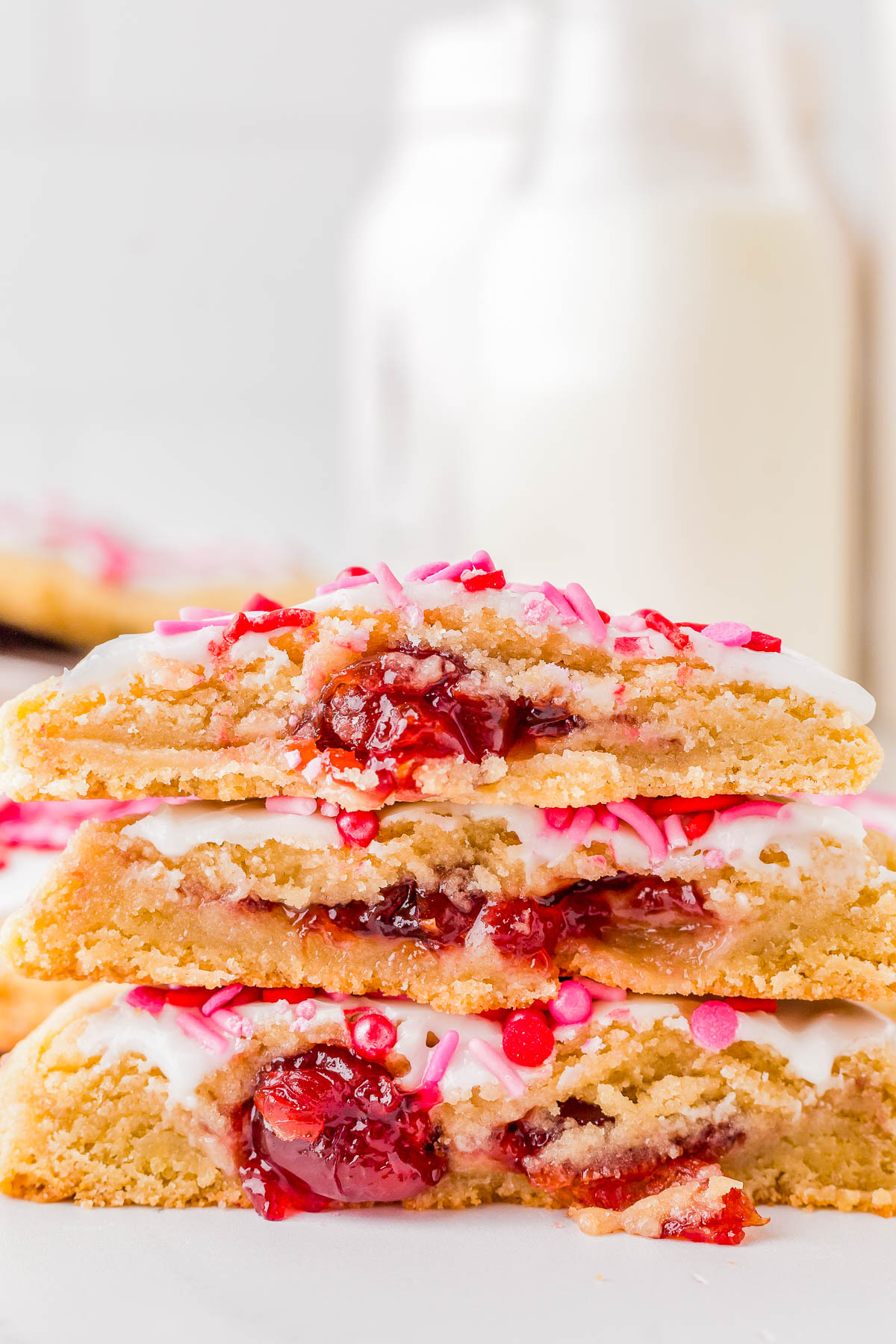 Stack of cookies with cherry filling, topped with white icing and pink sprinkles, with milk bottles in the background.