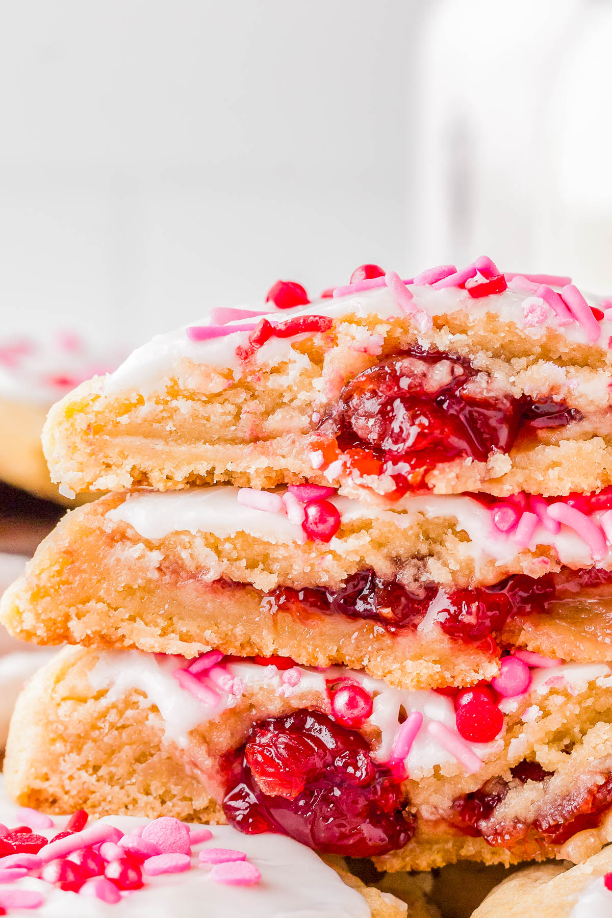 Close-up of stacked cookies filled with red berry jam, topped with white icing and pink sprinkles.