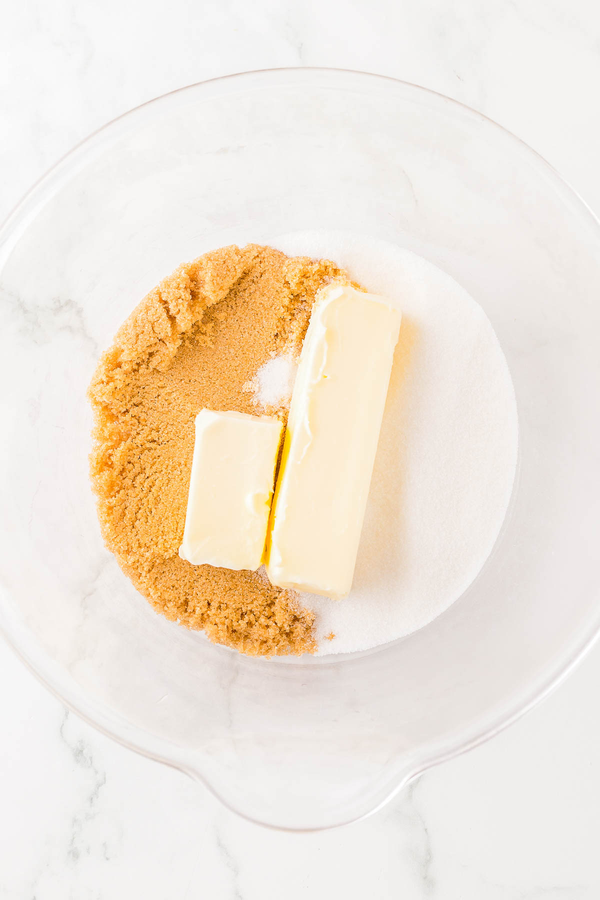 Brown sugar, white sugar, and two sticks of butter in a clear mixing bowl on a marble surface.