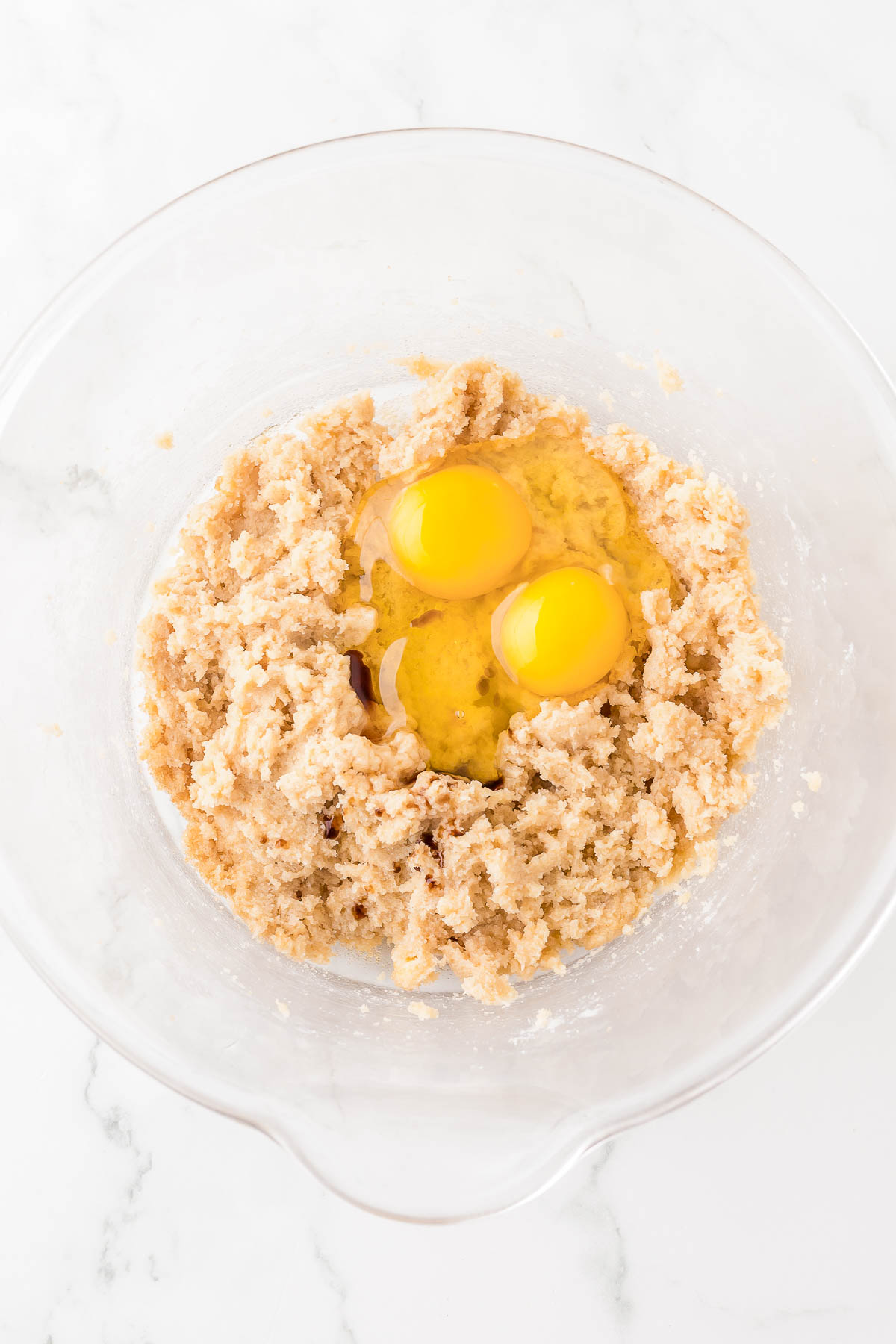 A glass bowl containing cookie dough with two cracked eggs in the center, on a white marble surface.