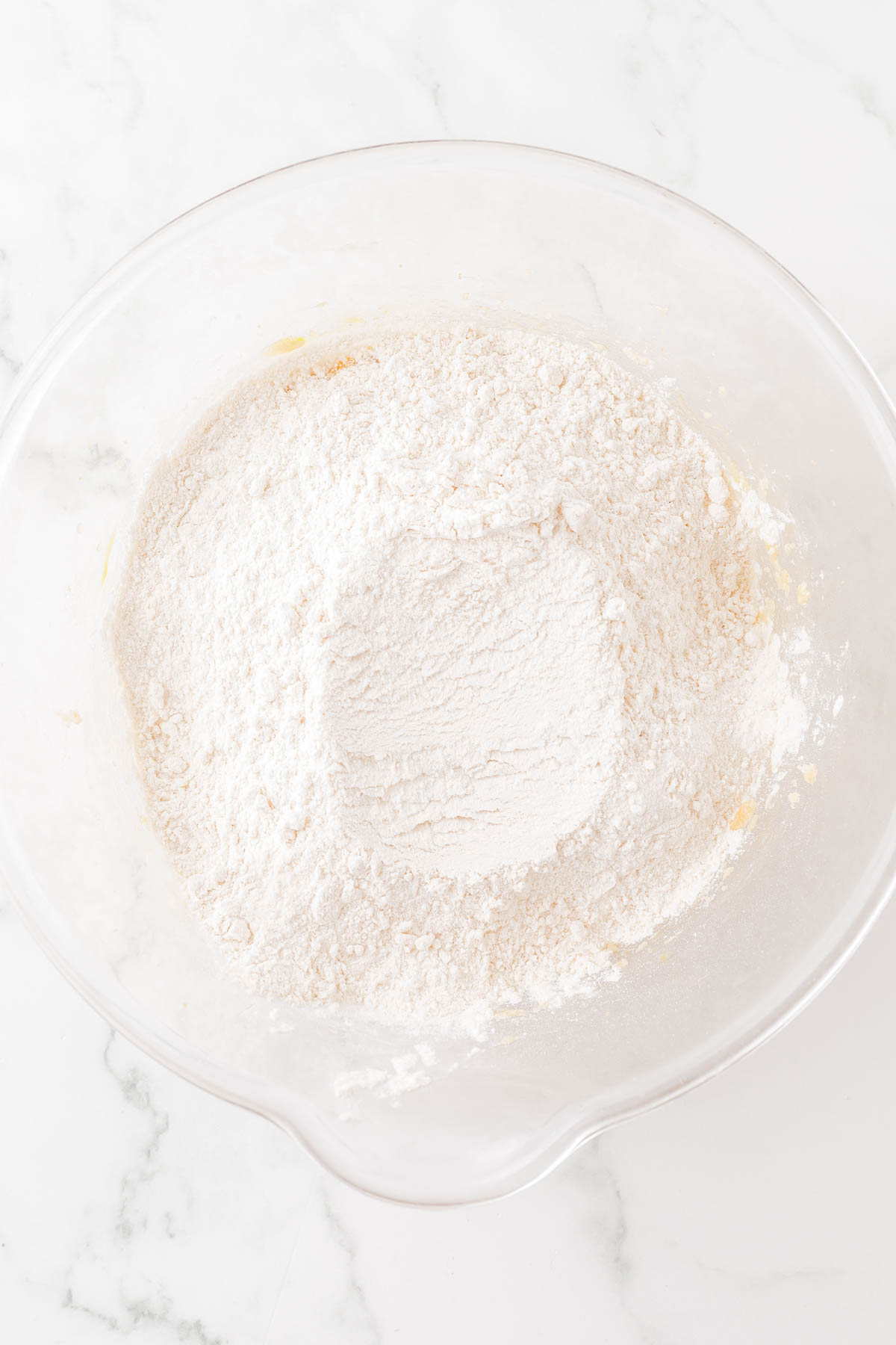 A glass bowl containing a mixture of flour and other dry ingredients on a white marble surface.