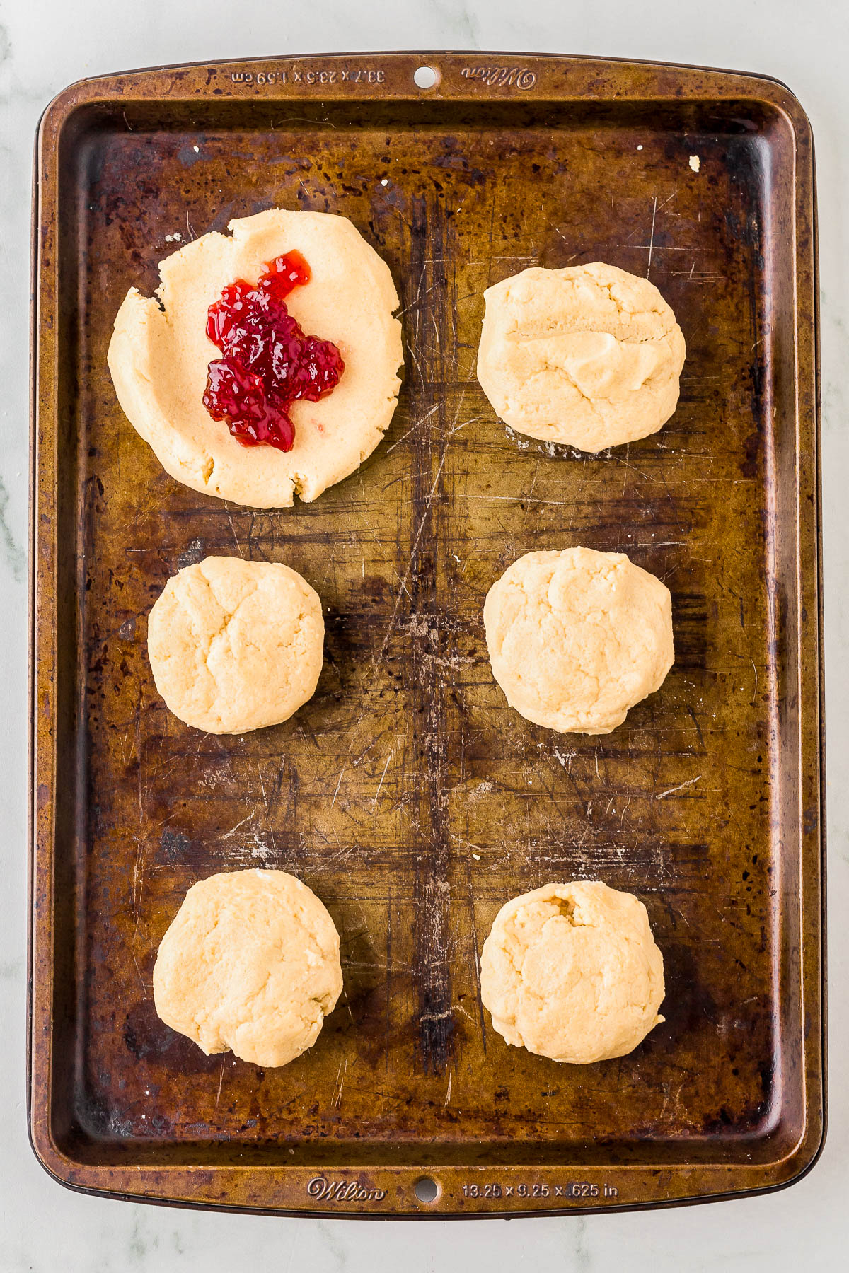 Six unbaked biscuits on a baking tray, with one biscuit partially spread with red jam.