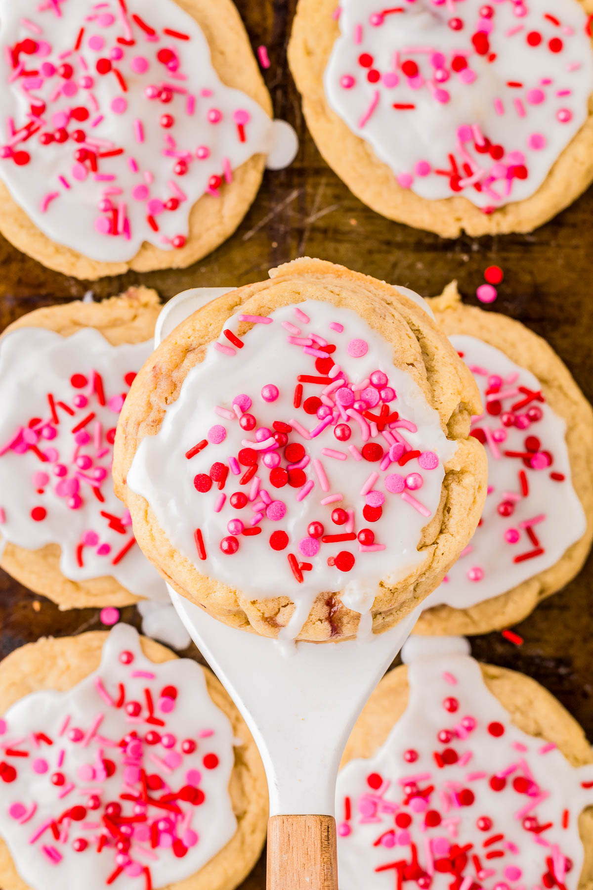 Cookies with white icing and red-pink sprinkles, one on a spatula above others on a baking sheet.