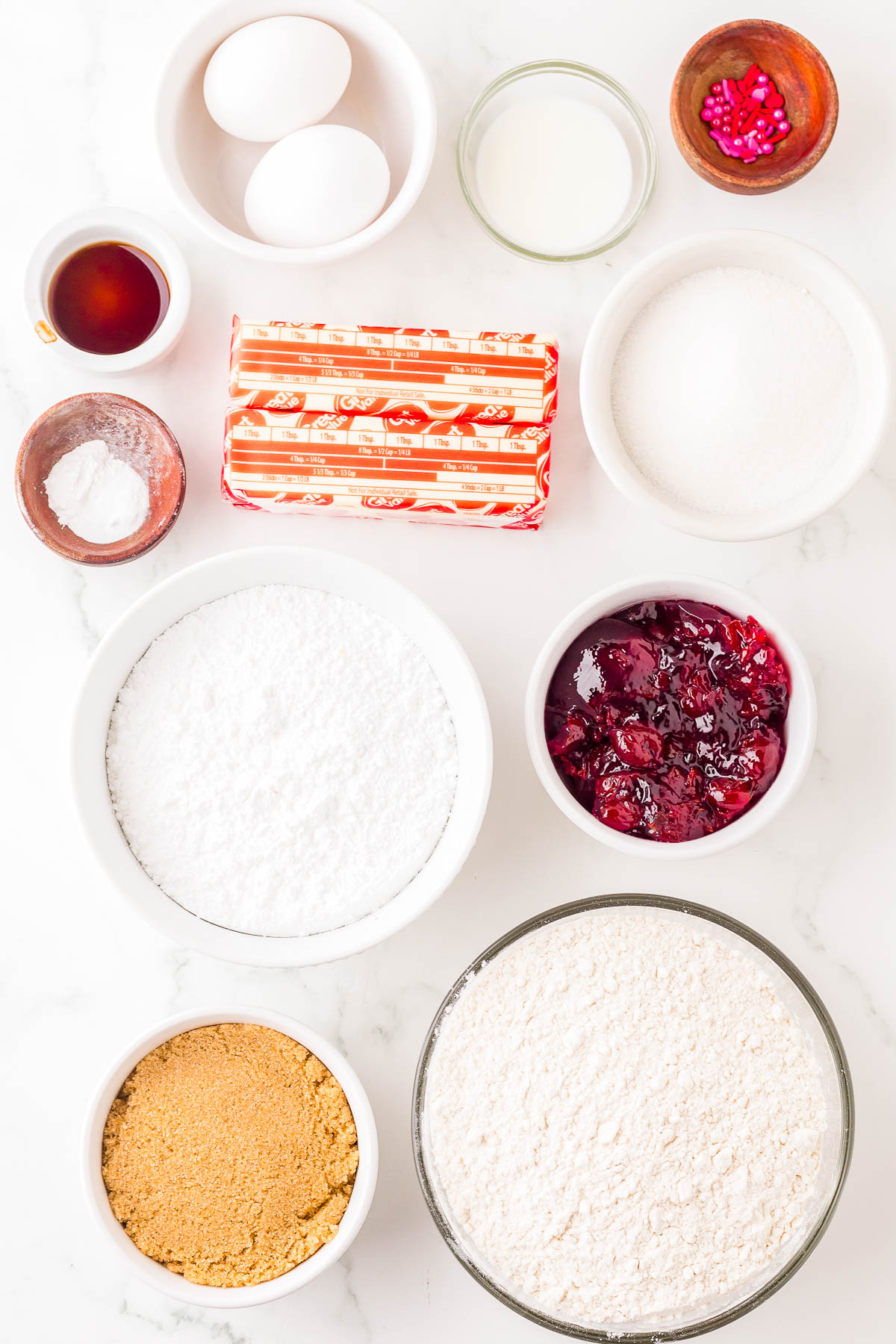An assortment of baking ingredients: eggs, butter, milk, cherries, sugar, brown sugar, flour, vanilla extract, powdered sugar, baking powder, and pink sprinkles in bowls on a marble surface.